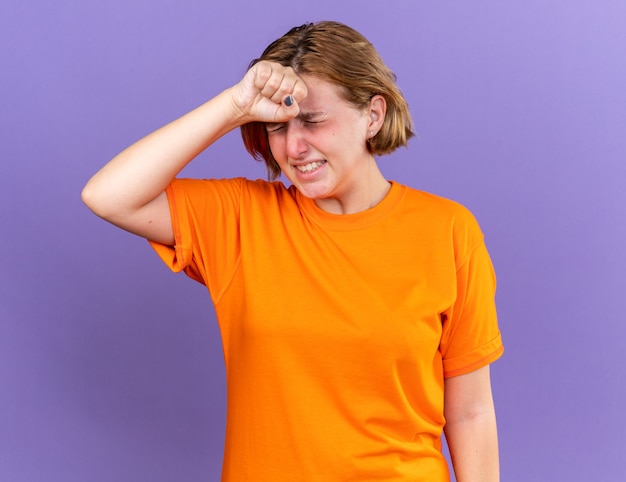 Ongezonde jonge vrouw in oranje t-shirt voelt zich vreselijk als ze haar voorhoofd aanraakt terwijl ze zich duizelig voelt met griep?