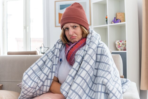 Ongezonde jonge vrouw in warme muts gewikkeld in deken die er onwel en ziek uitziet en lijdt aan verkoudheid en griep met thermometer die koorts heeft en er bezorgd uitziet zittend op de bank in een lichte woonkamer