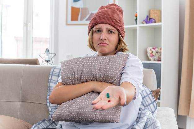 Ongezonde jonge vrouw in warme muts met deken die zich onwel en ziek voelt en lijdt aan verkoudheid en griep met kussen en pillen, bezorgd zittend op de bank in lichte woonkamer