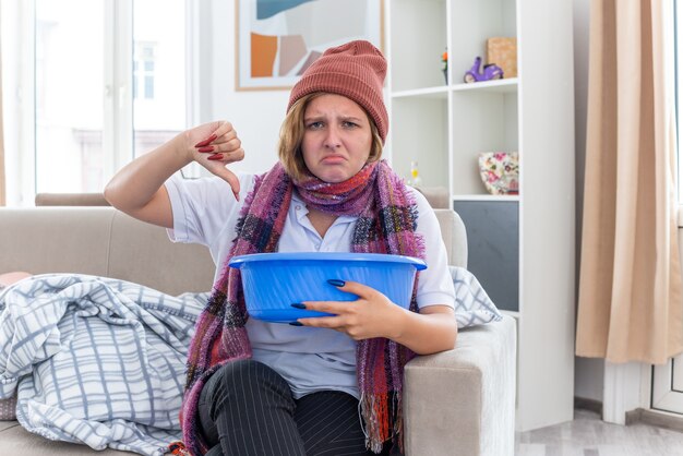 Ongezonde jonge vrouw in warme muts met sjaal om nek met bekken voelt zich misselijk en ziet er onwel en ziek uit met duimen naar beneden zittend op de bank in een lichte woonkamer