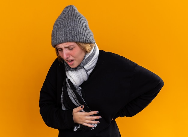 Ongezonde jonge vrouw met een warme muts en met sjaal om haar nek voelt zich ziek en lijdt aan griep die haar rug aanraakt met pijn die over de oranje muur staat