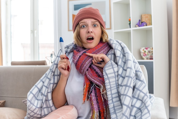 Ongezonde jonge vrouw met een warme muts gewikkeld in een deken die er onwel en ziek uitziet en lijdt aan verkoudheid en griep die een thermometer vasthoudt die koorts heeft en er bezorgd uitziet zittend op de bank in een lichte woonkamer