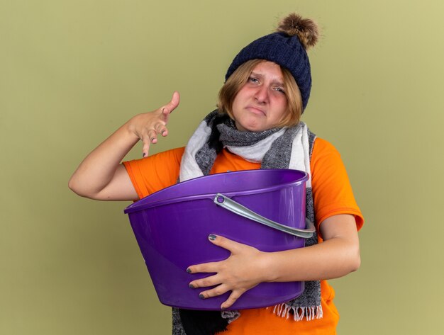 Ongezonde jonge vrouw met hoed met sjaal om haar nek voelt zich misselijk en heeft last van misselijkheid met een bekken dat over een groene muur staat