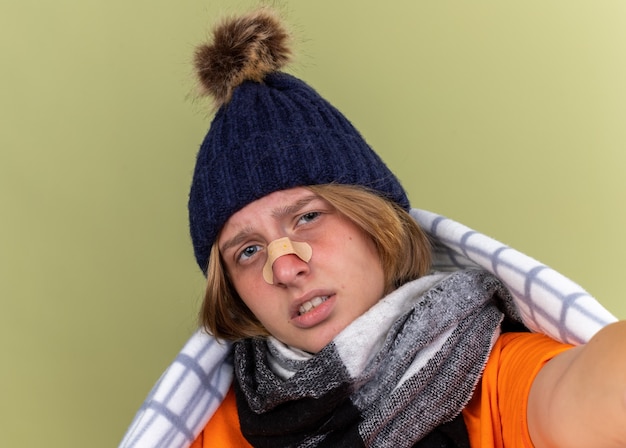 Gratis foto ongezonde jonge vrouw met warme muts en sjaal om nek gewikkeld in deken die lijdt aan kou met patch op haar neus met droevige uitdrukking die over groene muur staat
