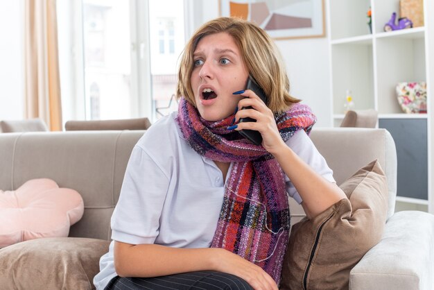 Ongezonde jonge vrouw met warme sjaal om nek onwel en ziek lijden aan griep en verkoudheid zittend op de Bank praten op mobiele telefoon in paniek zittend in lichte woonkamer