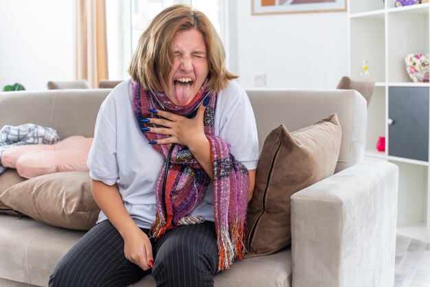 Gratis foto ongezonde jonge vrouw met warme sjaal om nek voelt zich verschrikkelijk, lijdt aan griep en verkoudheid, luistert naar haar longen met behulp van een stethoscoop hoesten in paniek zittend op de bank in een lichte woonkamer