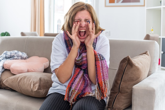 Gratis foto ongezonde jonge vrouw met warme sjaal om nek voelt zich vreselijk en lijdt aan virus dat in paniek schreeuwt terwijl ze op de bank zit in een lichte woonkamer