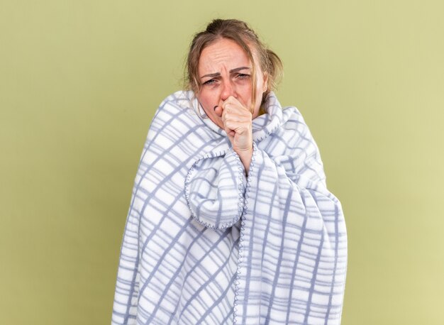 Ongezonde vrouw gewikkeld in een deken die zich vreselijk voelt en lijdt aan griep en koud hoesten die over een groene muur staat