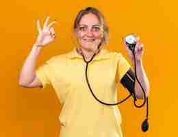 Gratis foto ongezonde vrouw in geel shirt die zich beter voelt om haar bloeddruk te meten met behulp van een tonometer die lacht met een ok-teken dat over de oranje muur staat