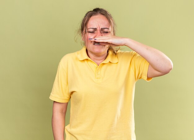 Ongezonde vrouw in geel shirt die zich vreselijk voelt, lijdt aan griep en verkoudheid die een loopneus afveegt