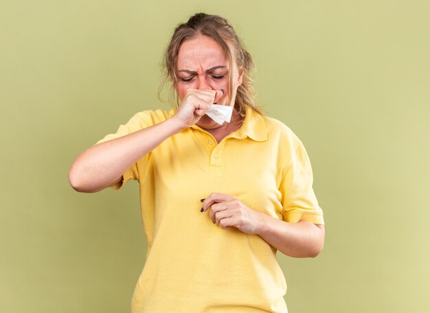 Ongezonde vrouw in geel shirt die zich vreselijk voelt, lijdt aan griep en verkoudheid met koorts die hoest over groene muur staat