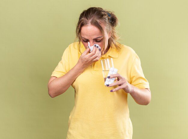 Ongezonde vrouw in geel shirt voelt zich verschrikkelijk met glas water en pillen die lopende neus snuiten niezen in weefsel dat lijdt aan griep die over groene muur staat