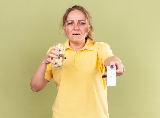 Ongezonde vrouw in geel shirt voelt zich vreselijk met glas water en pillen die aan griep lijden