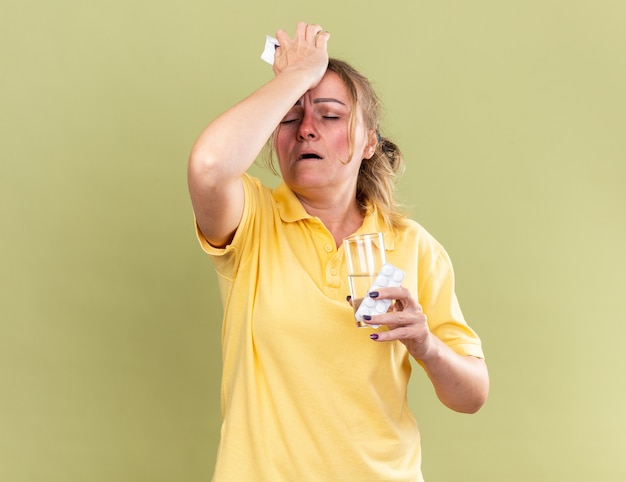 Ongezonde vrouw in geel shirt voelt zich vreselijk met glas water en pillen die haar voorhoofd aanraken en lijdt aan sterke hoofdpijn vanwege een verstopte neus met griep