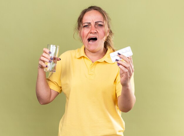 Ongezonde vrouw in geel shirt voelt zich vreselijk met glas water en pillen die lijden aan griep en verkoudheid over groene muur staan