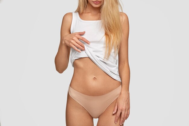 Onherkenbare blonde dame toont een platte buik en een slanke taille, tilt een casual wit vest op, draagt een broek, toont een perfect slank lichaam, staat tegen de muur. Mensen, gezonde levensstijl concept