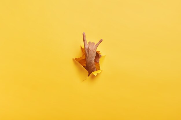 Onherkenbare man maakt rock n roll-gebaar door gescheurd gat in geel papier. Mannetje toont hoorn bord met hand gestrekt in de opening van het papier. Lichaamstaal concept. Gekleurde ruimte
