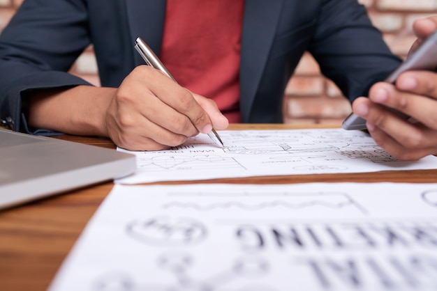 Gratis foto onherkenbare man zit aan tafel met smartphone en businessdiagram tekenen op papier