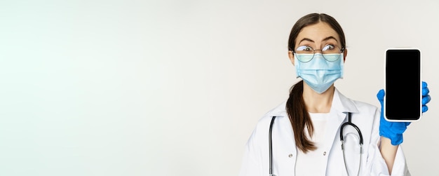 Online medisch hulpconcept verraste vrouwelijke arts met gezichtsmasker die verbaasd keek terwijl hij mobiel liet zien