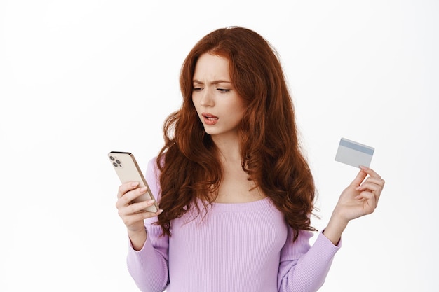 Online winkelen. Afbeelding van verward en overstuur roodharig meisje dat naar haar bankrekening op mobiele telefoon kijkt, teleurgesteld naar het scherm van de smartphone staart, creditcard vasthoudt, witte achtergrond