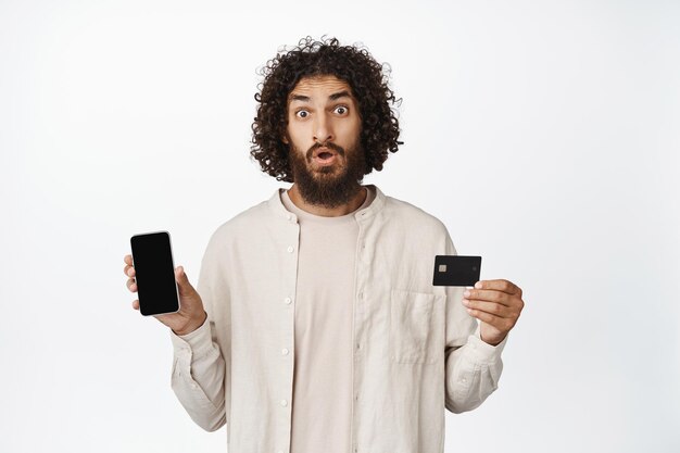 Online winkelen Jonge verbaasde Arabische man met het scherm van de mobiele telefoon en creditcard die onder de indruk van de camera op de witte achtergrond staart