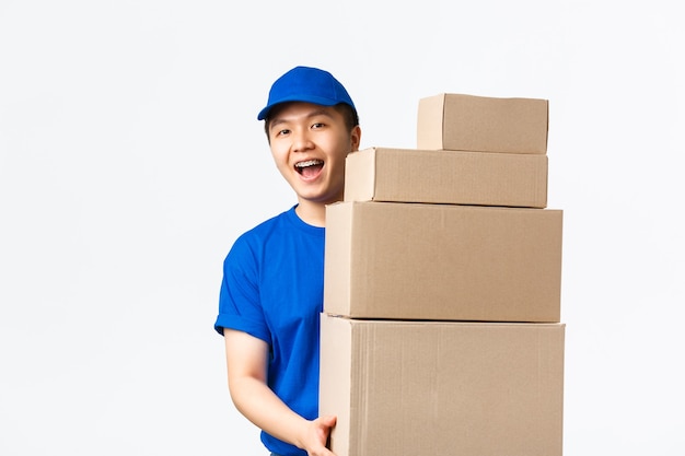 Online winkelen, snel verzendconcept. Vriendelijk lachende jonge aziatische mannelijke koerier in blauw uniform draagt dozen met bestellingen. Bezorger brengt pakketten naar uw deur, staande witte achtergrond.
