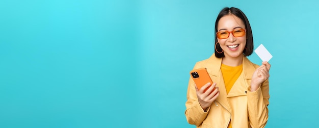 Online winkelen stijlvolle jonge aziatische vrouw in zonnebril die creditcard toont en smartphone gebruikt die op internet betaalt en een aankoop doet die over blauwe achtergrond staat