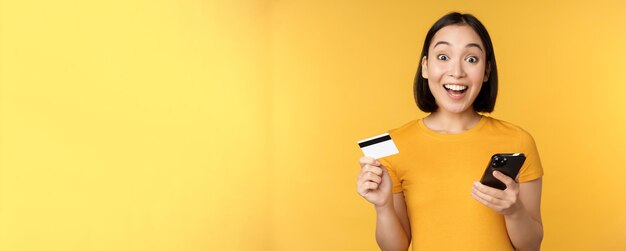Online winkelen Vrolijk Aziatisch meisje met creditcard en smartphone die de bestelling betaalt met een mobiele telefoon die op een gele achtergrond staat