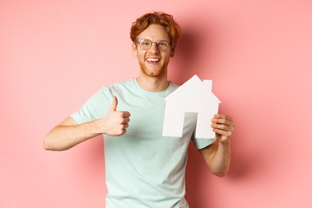 Onroerend goed. Vrolijke man in glazen en t-shirt die makelaarsbureau aanbeveelt, papier huis uitgesneden en thumbs-up toont, staande over roze achtergrond.