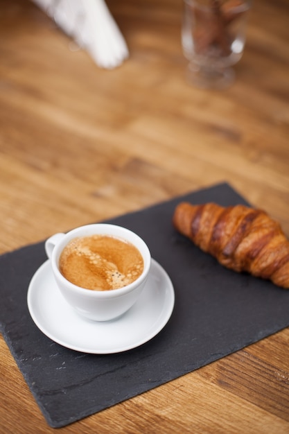 Ontbijt met espressokopje warme koffie en croissant op een zwarte stenen plaat. Koffie aroma.