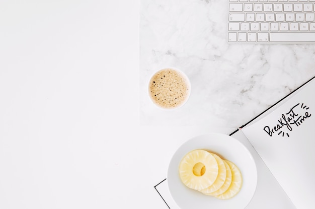 Ontbijttijd tekst op papier met toetsenbord; ananas plakjes en koffiekopje op het bureau