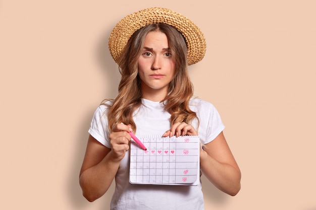 Ontevreden blanke vrouw in casual t-shirt en strooien hoed, geeft aan op periodekalender, wil geen menstruatie hebben tijdens rust aan zee, geïsoleerd op beige. Ongelukkige vrouw binnen