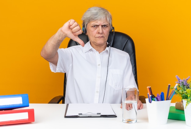 Ontevreden blanke vrouwelijke callcenter-operator op koptelefoon zittend aan bureau met kantoorhulpmiddelen die naar beneden wijzen geïsoleerd op oranje muur