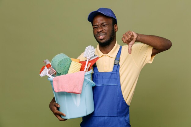 Ontevreden duimen naar beneden houden emmer schoonmaak tools jonge Afro-Amerikaanse schonere man in uniform met handschoenen geïsoleerd op groene achtergrond