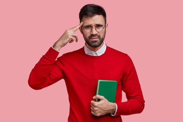 Ontevreden Europese man houdt de vinger op de tempel, draagt een rode trui, draagt een groen leerboek, heeft een ongelukkige gezichtsuitdrukking