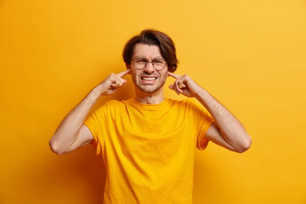 ontevreden Europese man klemt tanden niet scherp vanwege lawaai draagt bril casual t-shirt hoort hartverscheurende schreeuw geïsoleerd over gele muur. Niet willen luisteren