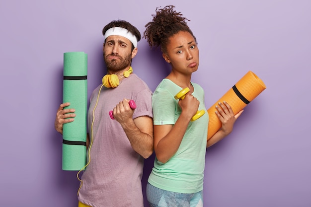 Gratis foto ontevreden gemengd ras paar staan tegen elkaar, heffen zware halters op, voelen zich moe na actieve sporttraining in de sportschool, gebruiken karemat