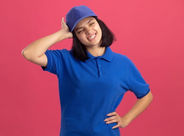 Ontevreden jong leveringsmeisje in blauw uniform en pet die haar hoofd aanraken met hoofdpijn die zich over roze muur bevindt