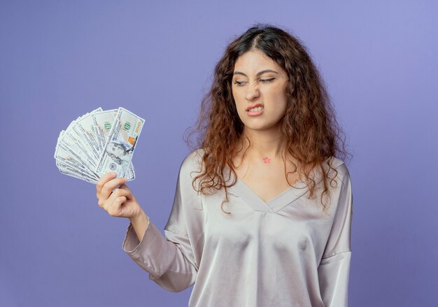 Ontevreden jong mooi meisje dat en contant geld houdt bekijkt dat op blauwe muur wordt geïsoleerd