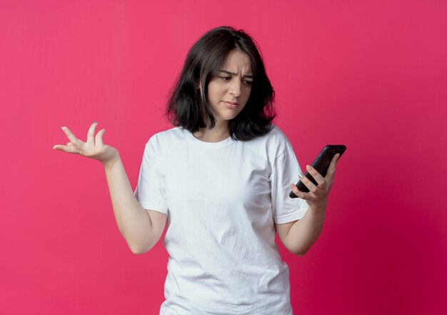 Ontevreden jong vrij Kaukasisch meisje dat en mobiele telefoon houdt bekijkt en lege die hand op karmozijnrode achtergrond met exemplaarruimte toont