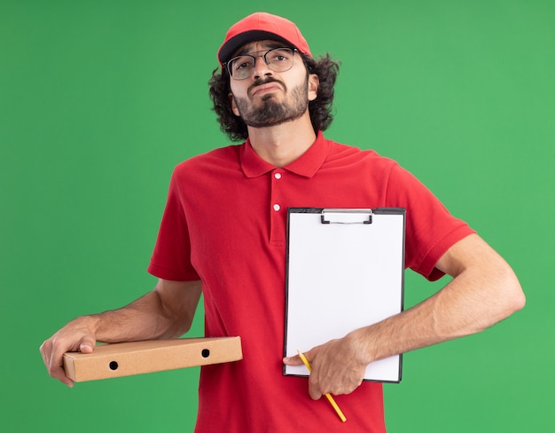 Ontevreden jonge blanke bezorger in rood uniform en pet met bril met pizzapakket potlood met klembord naar camera geïsoleerd op groene muur