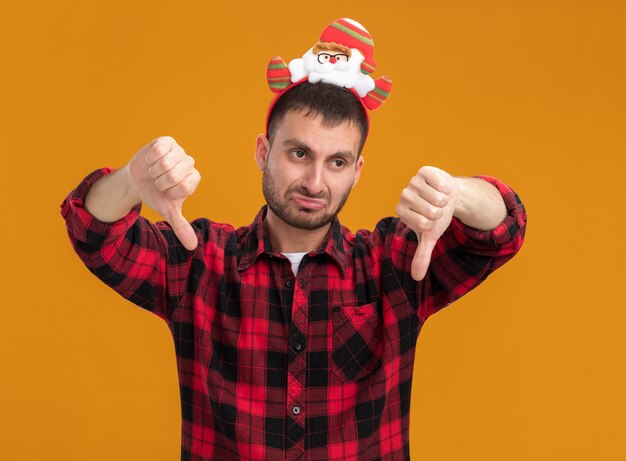 Ontevreden jonge blanke man met de hoofdband van de kerstman kijken naar kant tonen duimen omlaag geïsoleerd op een oranje achtergrond