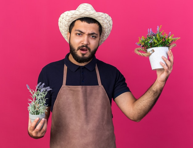 ontevreden jonge blanke mannelijke tuinman die tuinieren hoed draagt ?? die bloempotten houdt die op roze muur met exemplaarruimte worden geïsoleerd