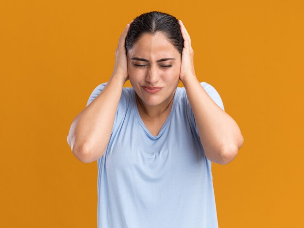 Ontevreden jonge brunette blanke meid bedekt oren met handen geïsoleerd op een oranje muur met kopieerruimte