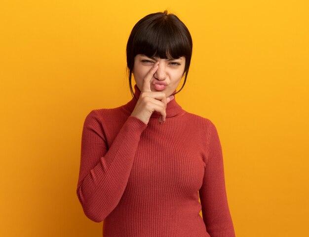 Ontevreden jonge brunette blanke meid legt vinger op neus geïsoleerd op oranje muur met kopieerruimte