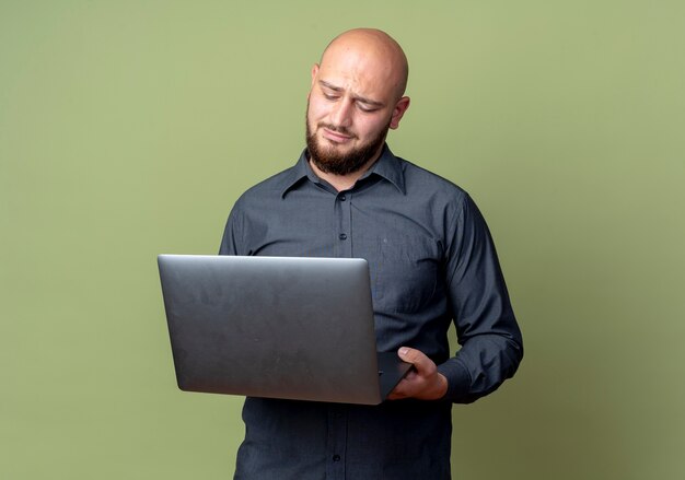 Ontevreden jonge kale call center man houden en kijken naar laptop geïsoleerd op olijfgroene achtergrond met kopie ruimte