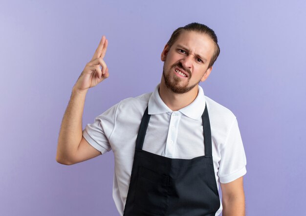 Gratis foto ontevreden jonge knappe kapper die eenvormig draagt die twee met hand toont die op purpere achtergrond wordt geïsoleerd