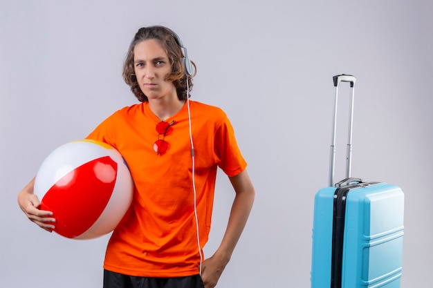 Ontevreden jonge knappe kerel in oranje t-shirt met hoofdtelefoons die opblaasbare bal houden die zich dichtbij reiskoffer bevindt