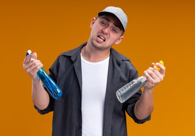 Ontevreden, jonge, knappe schoonmaakster met een T-shirt en dop met spuitflessen geïsoleerd op een oranje muur orange