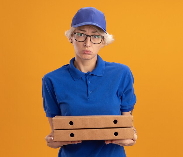 Ontevreden jonge leveringsvrouw in blauw uniform en pet die glazen dragen die pizzadozen houden die met droevige uitdrukking over oranje muur kijken